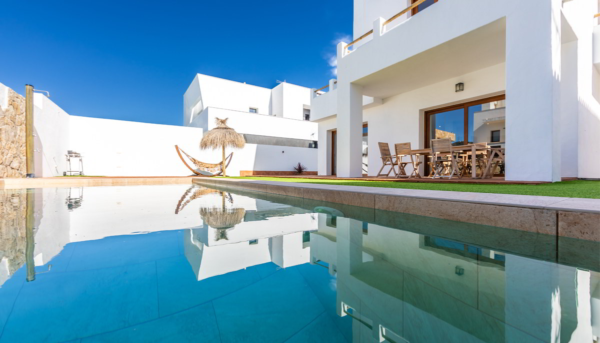 Piscina privada con reflejo de vivienda blanca y cielo azul.