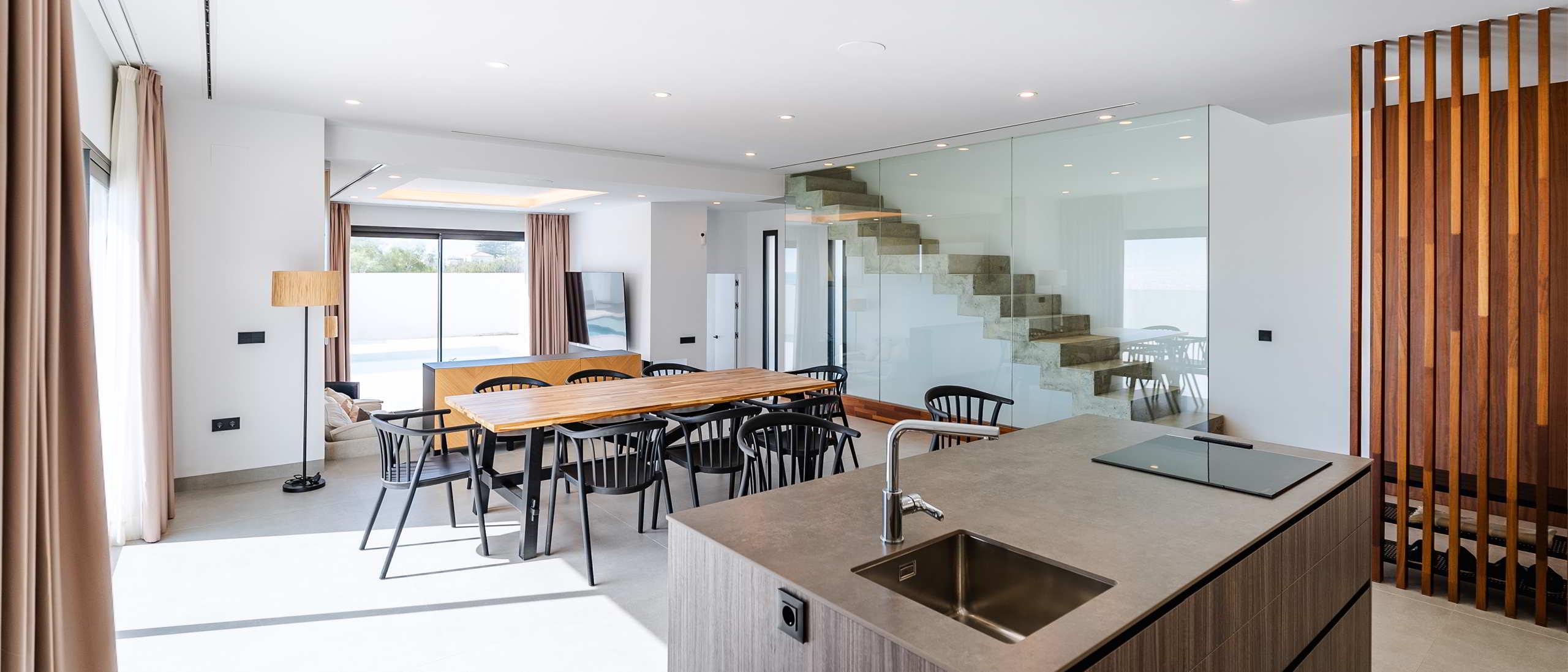 Espacio diáfano con cocina abierta, comedor y escalera de hormigón visto con cerramiento de vidrio.