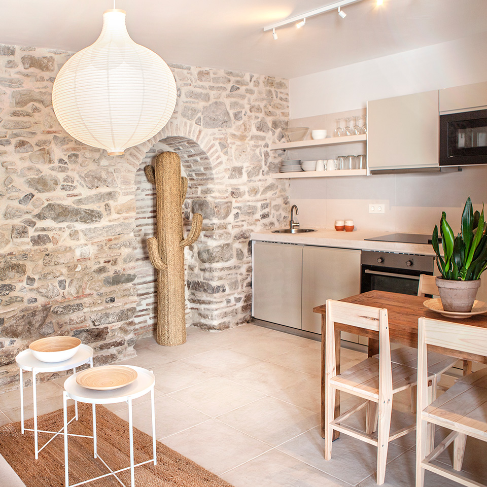 Cocina abierta con muebles de madera clara, mesa y sillas de comedor de madera y pared de piedra a vista