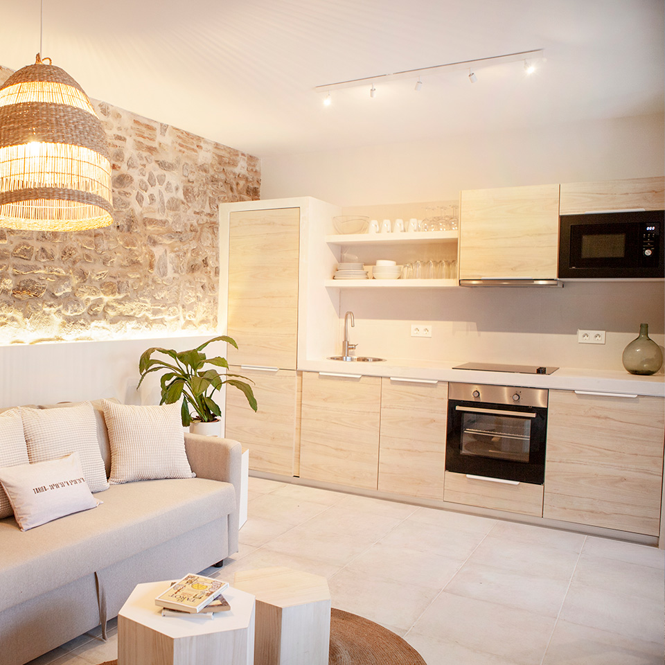 Cocina abierta con muebles de madera clara, salón y pared de piedra a vista