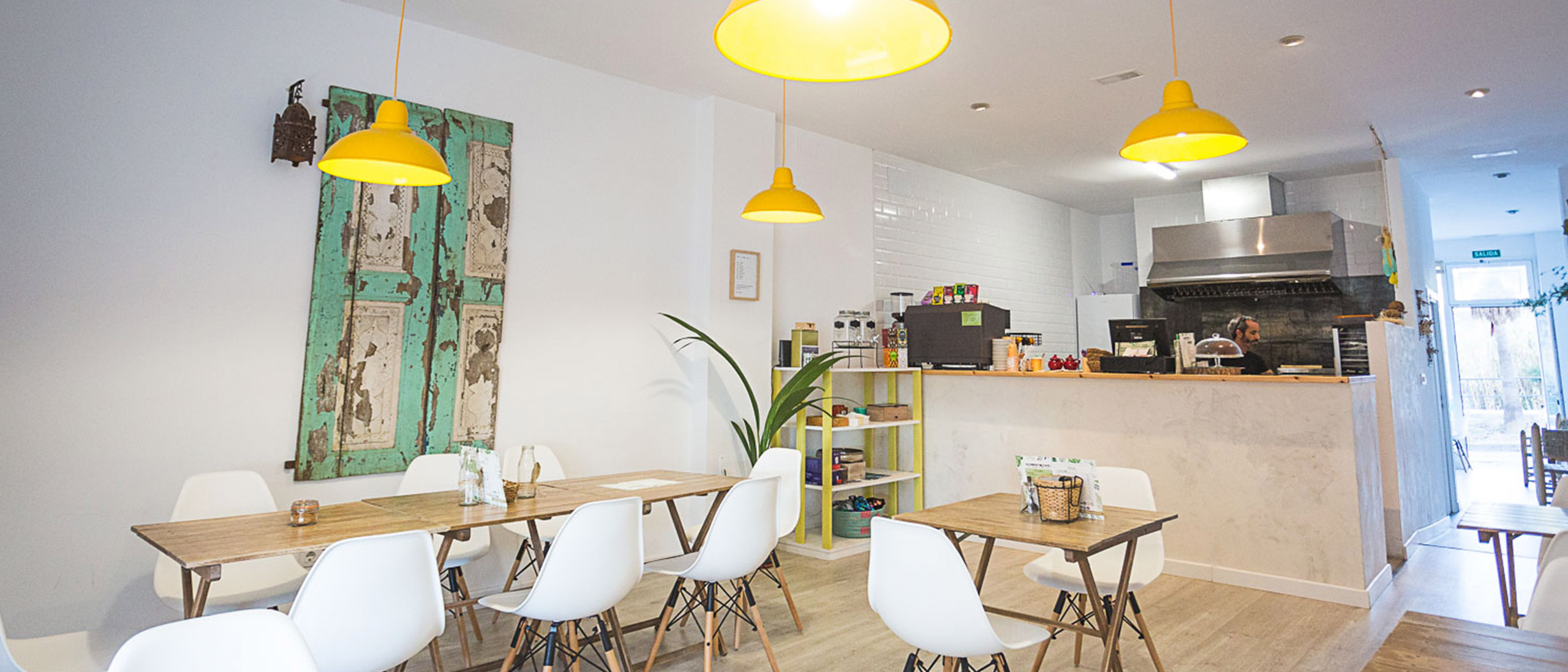 Interior del Café con cocina avista y salón con mesas de madera y sillas blancas