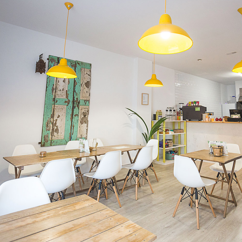 Interior del Café con cocina a vista y salón con mesas de madera y sillas blancas